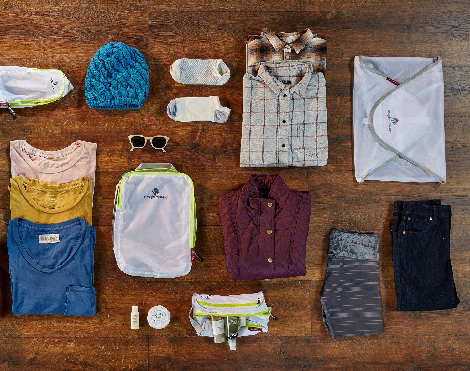 Draufsicht auf Holztisch mit Pack-It Cubes von Eagle Creek, T-Shirts und Reiseaccessoires